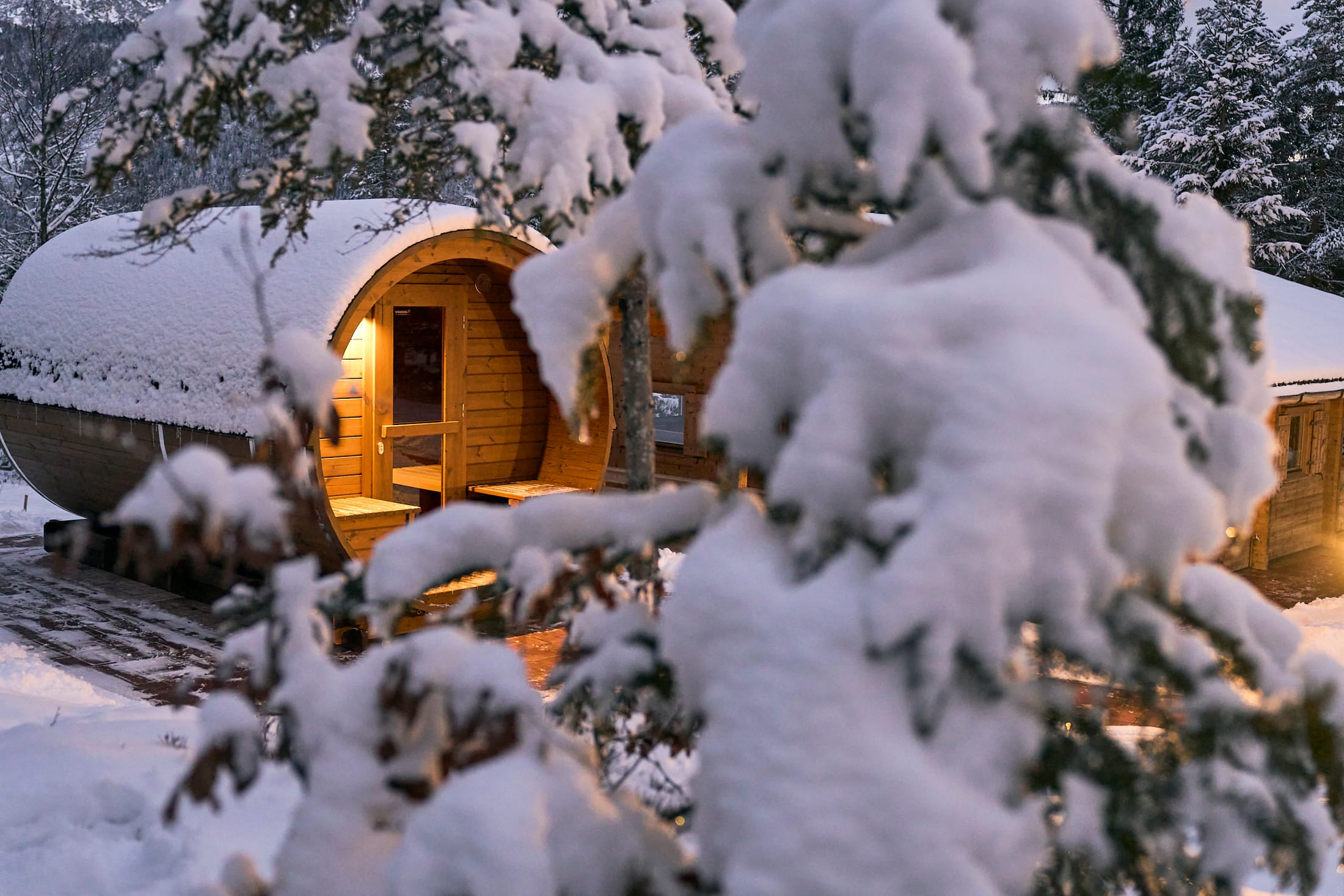 Warm beleuchtete Fass-Sauna im verschneiten Wald bei Dämmerung